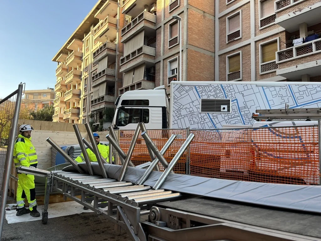 Operatori in alta visibilità accanto a una rulliera con materiale tubolare grigio, in un cantiere stradale davanti a un camion SAPI.