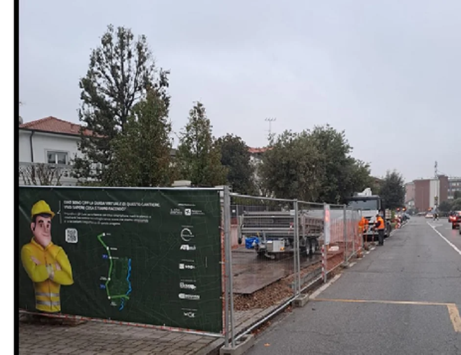 Cantiere lungo una strada urbana, delimitato da transenne con un pannello informativo e mezzi di lavoro.