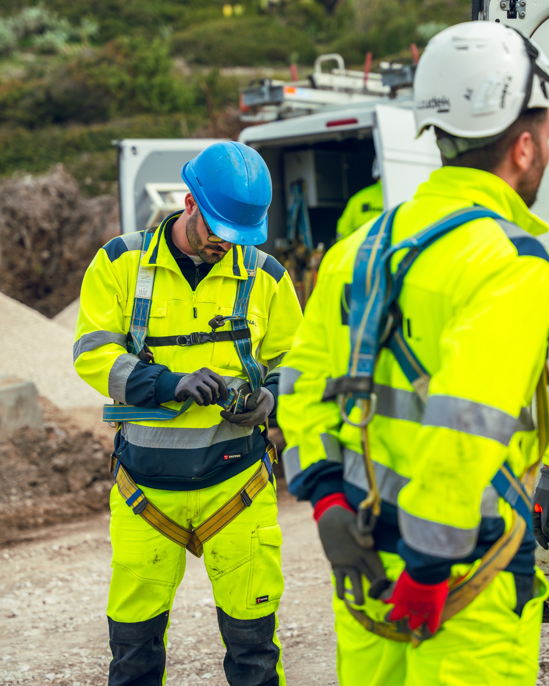 Operatore con casco blu e guanti che regola l’imbracatura, accanto a un collega in alta visibilità