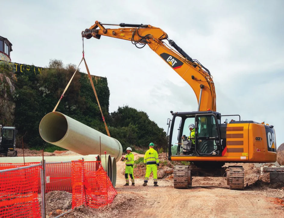 Escavatore arancione che sostiene una sezione di tubo con cinghie, con operatori in alta visibilità e recinzioni di cantiere.