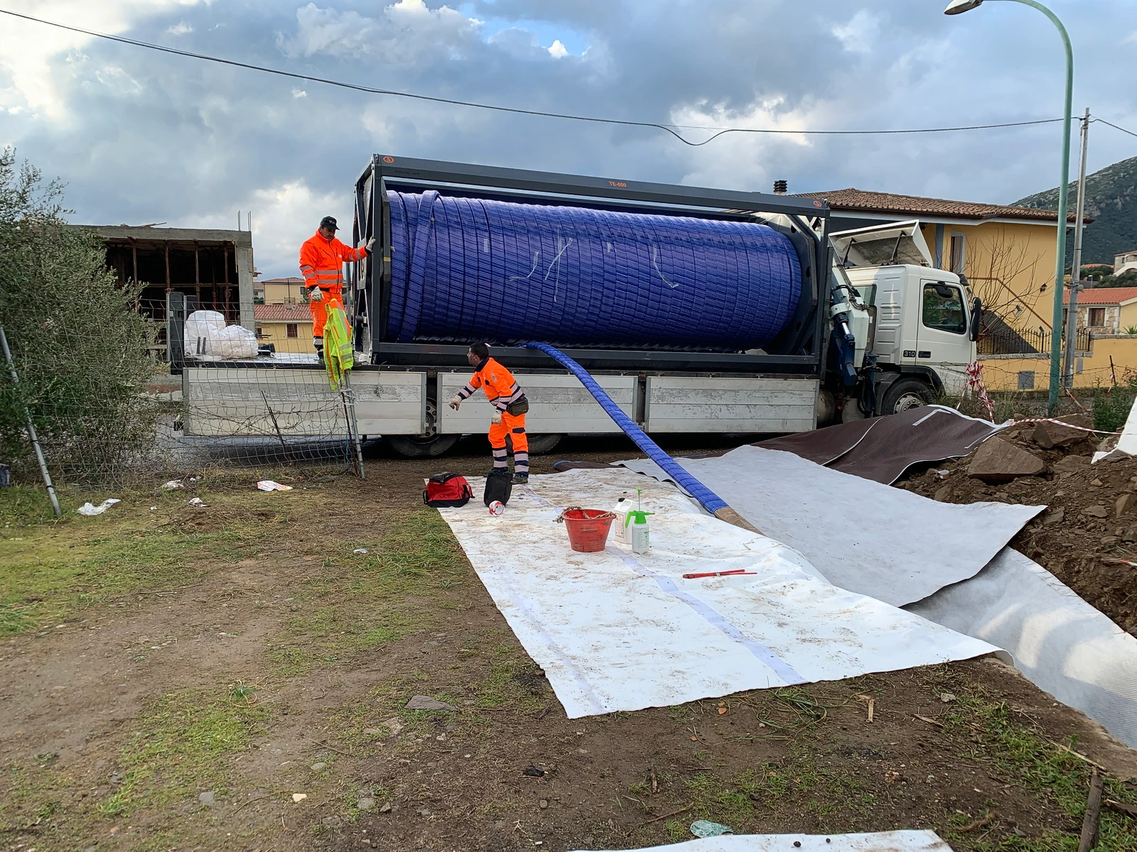 Bobina di liner blu sul camion e operatori accanto ai teli stesi verso lo scavo