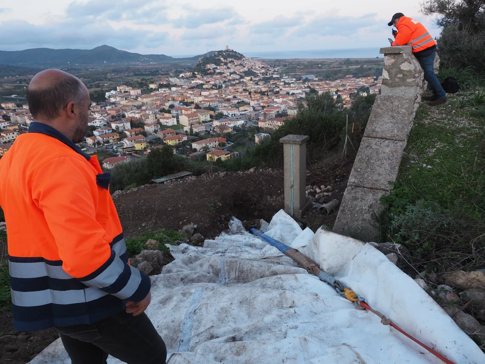 Due operatori in arancione osservano una linea tubolare posata su teli bianchi lungo un pendio, con abitato e costa sullo sfondo.